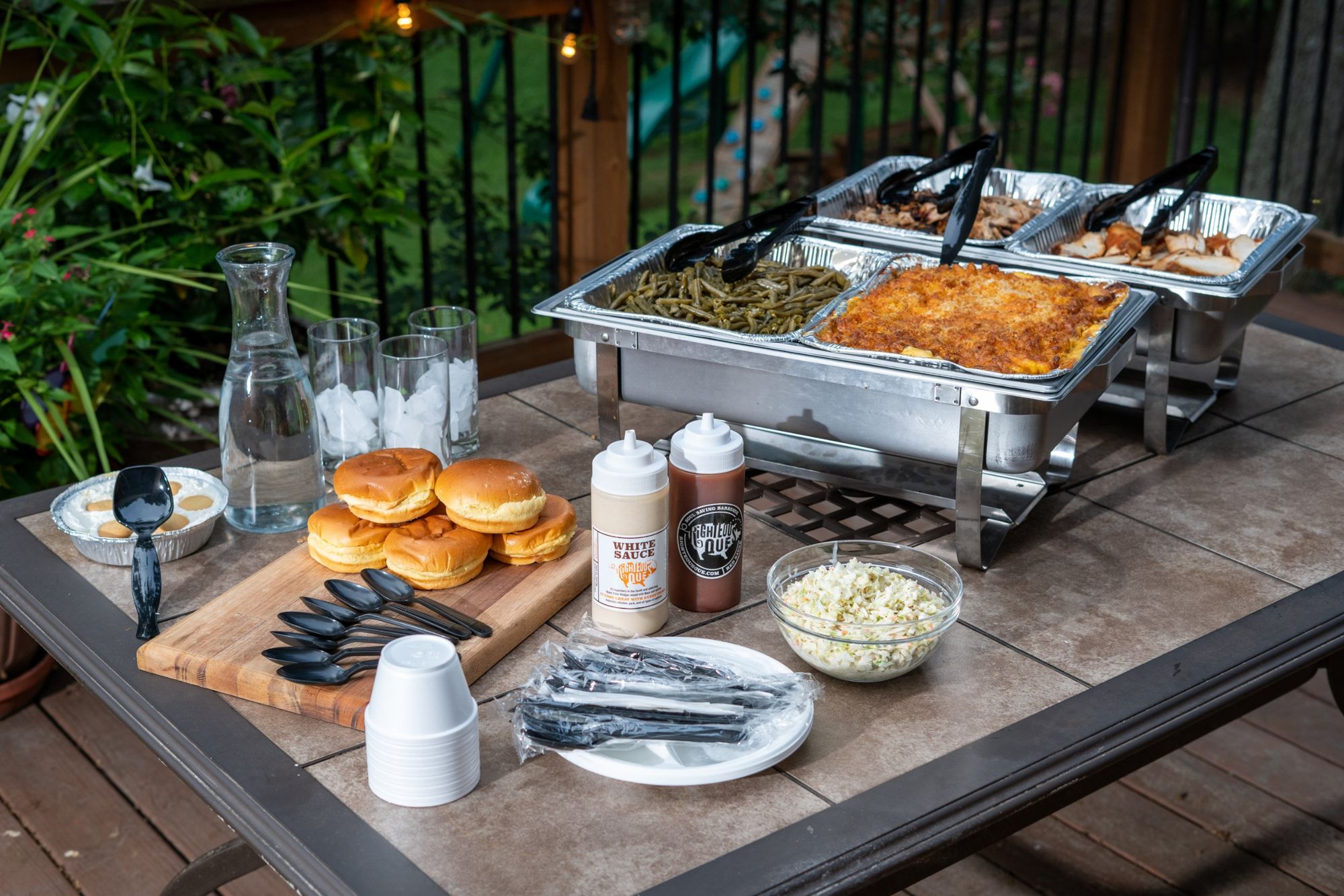 A Righteous 'Que catering spread on a backyard deck with chafing dishes, buns, sauces and slaw