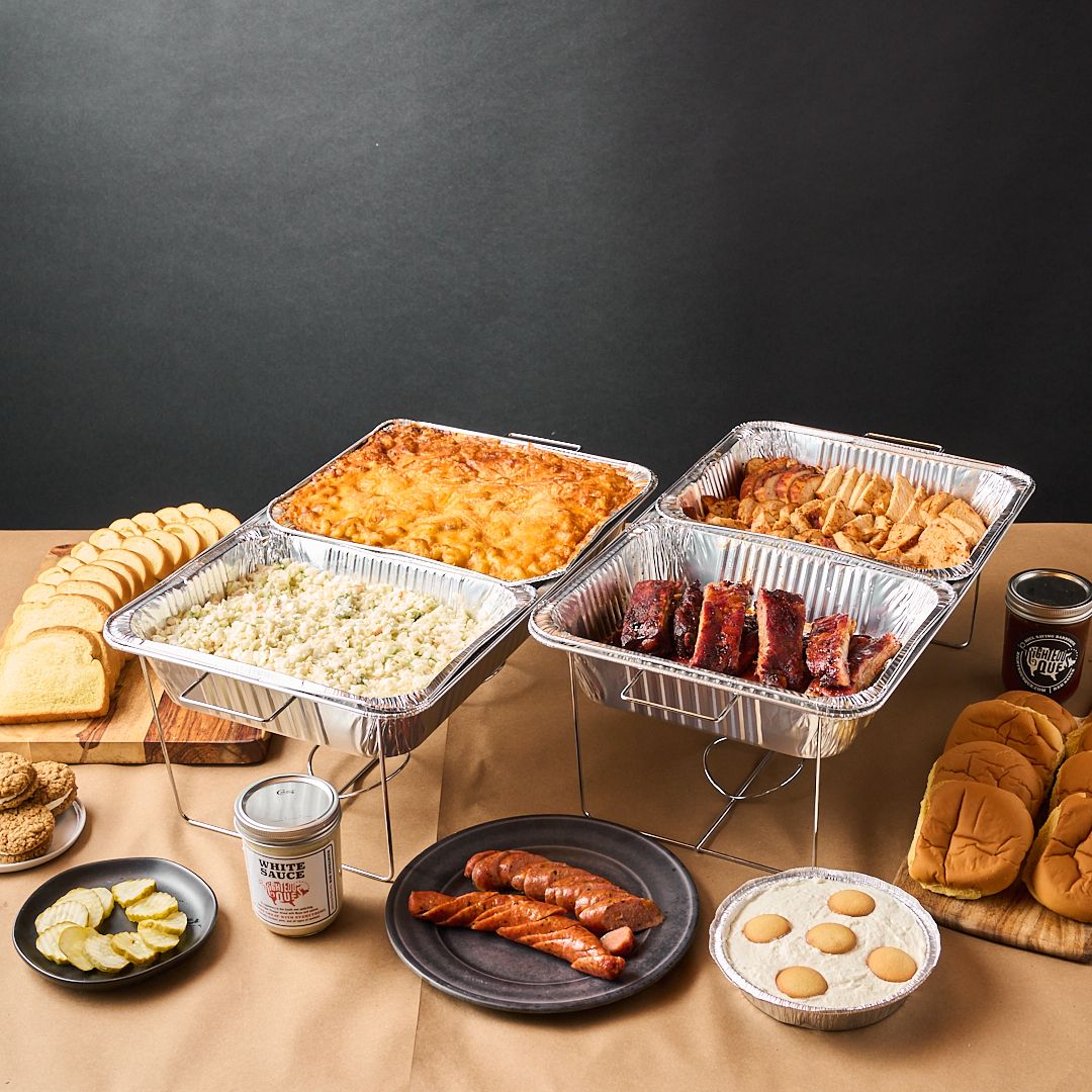 A full catering spread with chafing trays, ribs, chicken, sausage, buns and banana pudding