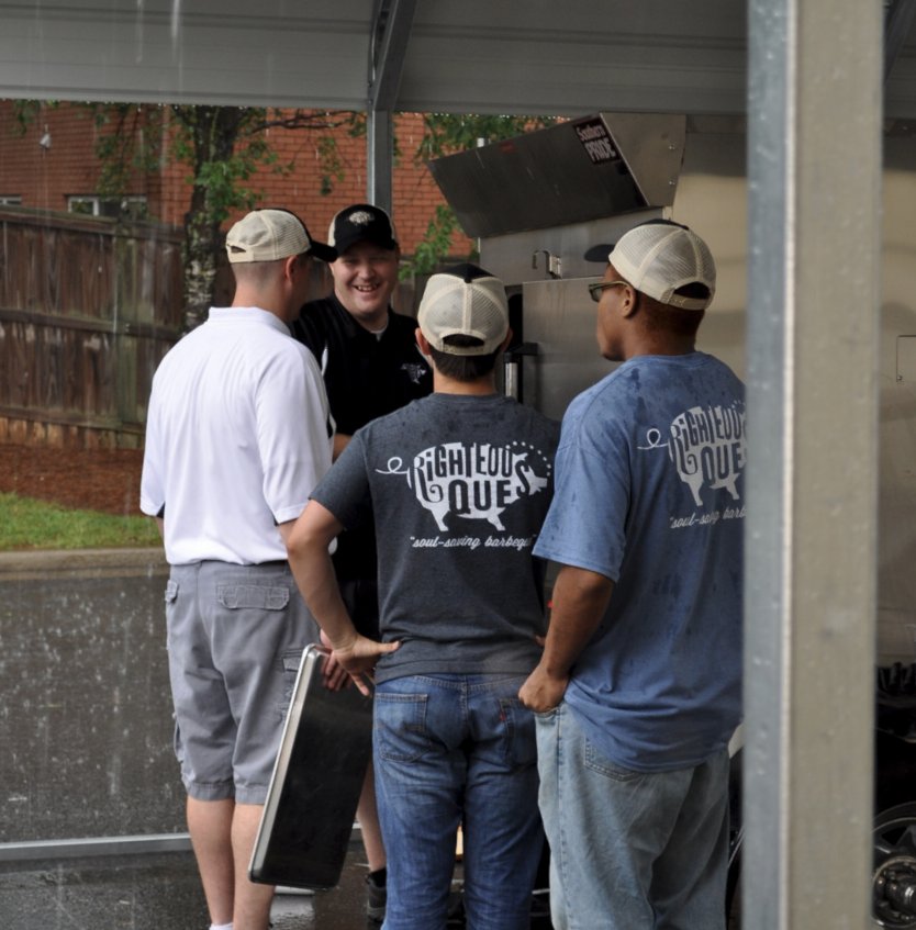 The Righteous Que crew in branded shirts gathered around the smoker